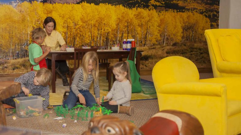 Children playing on carpeted floor and adult sitting at table helping a child paint with brush