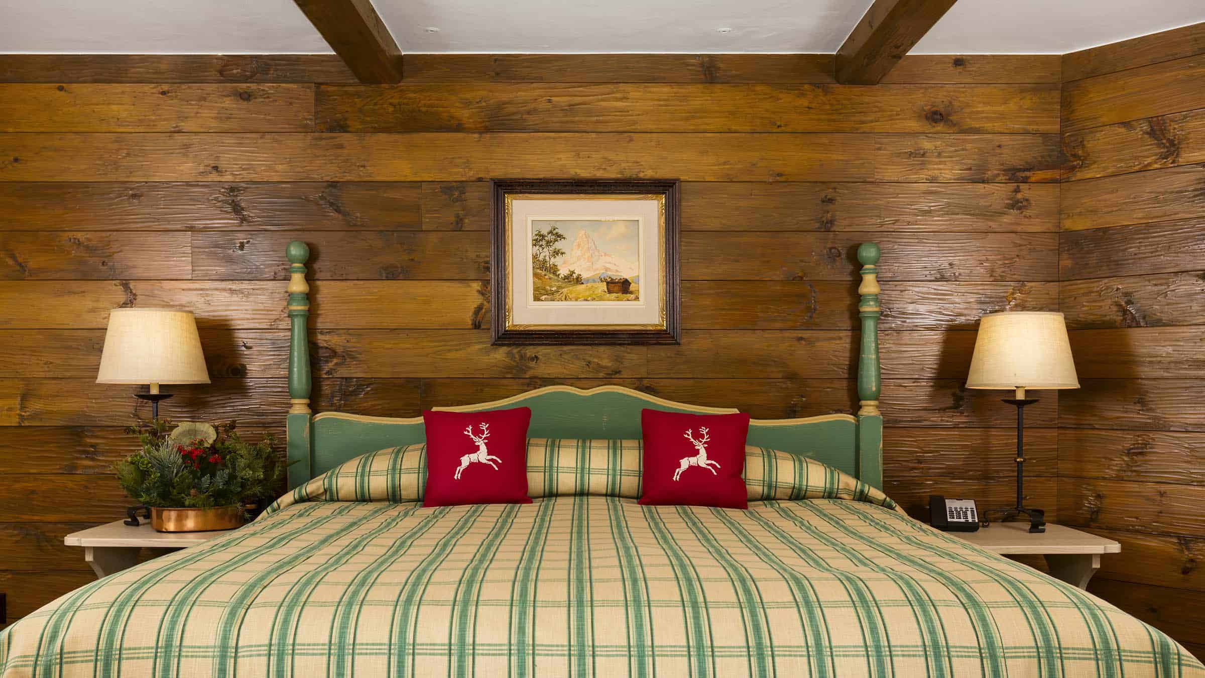 A bed with a green and beige plaid comforter, two red pillows with white deer, and a wooden headboard against a wood-paneled wall.