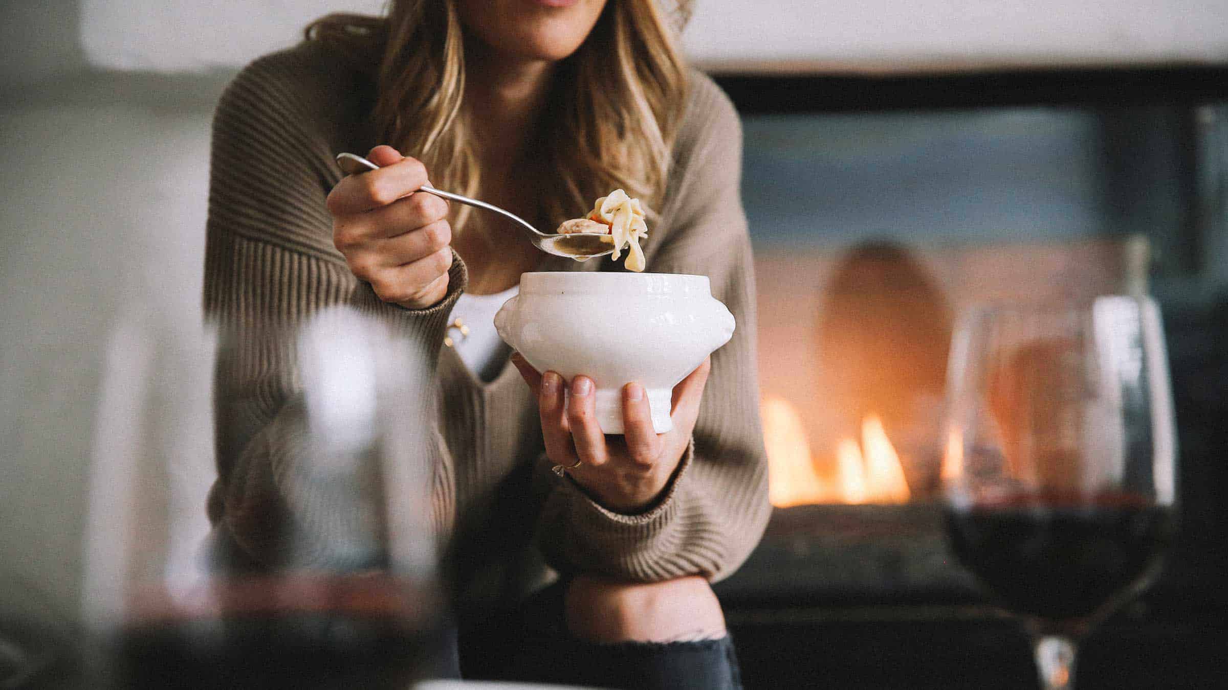 Woman eating noodle soup from a bowl, fireplace in the background