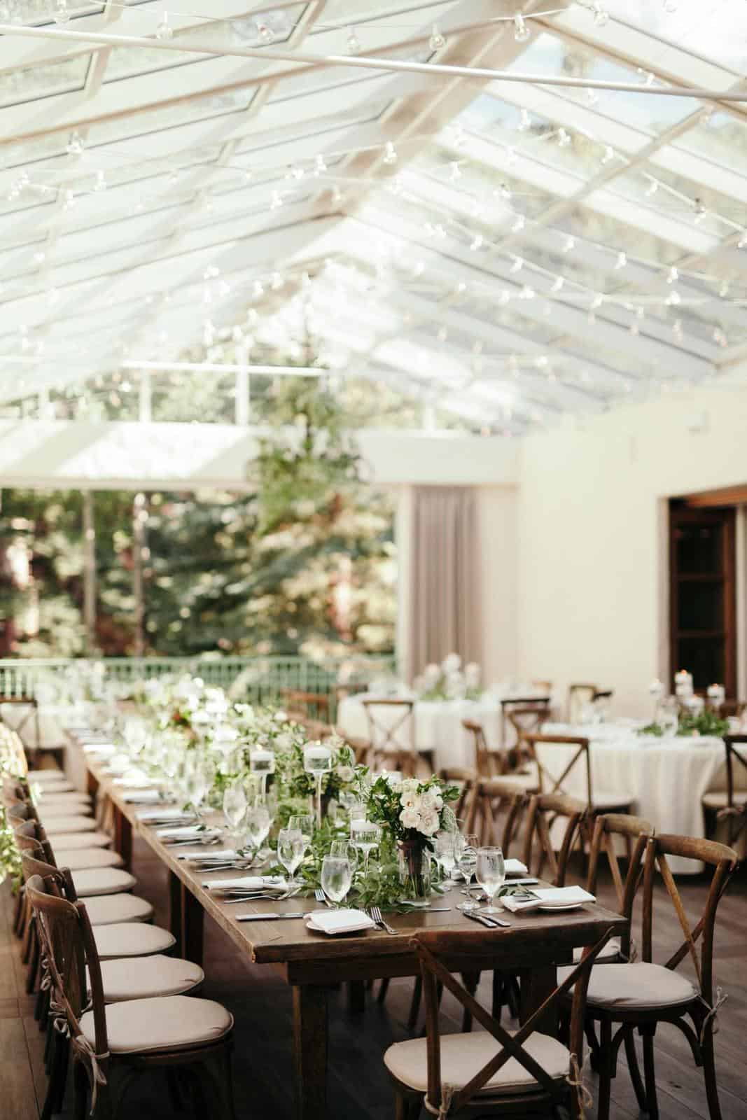 a long wooden table set with white flowers, glassware, and wooden chairs under string lights in a bright room
