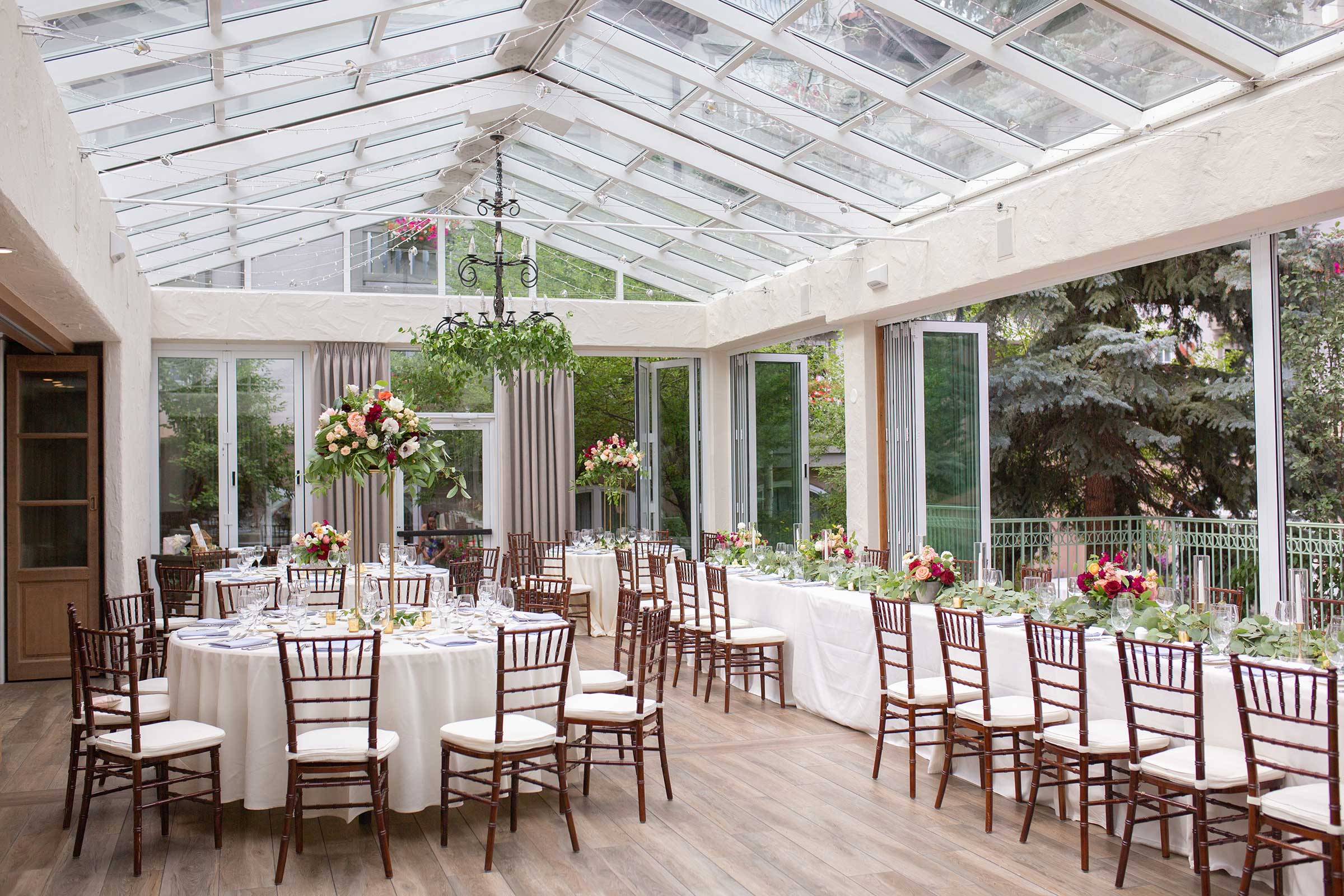 A brightly lit, elegantly set dining room with round tables, white tablecloths, and floral arrangements under a glass roof.