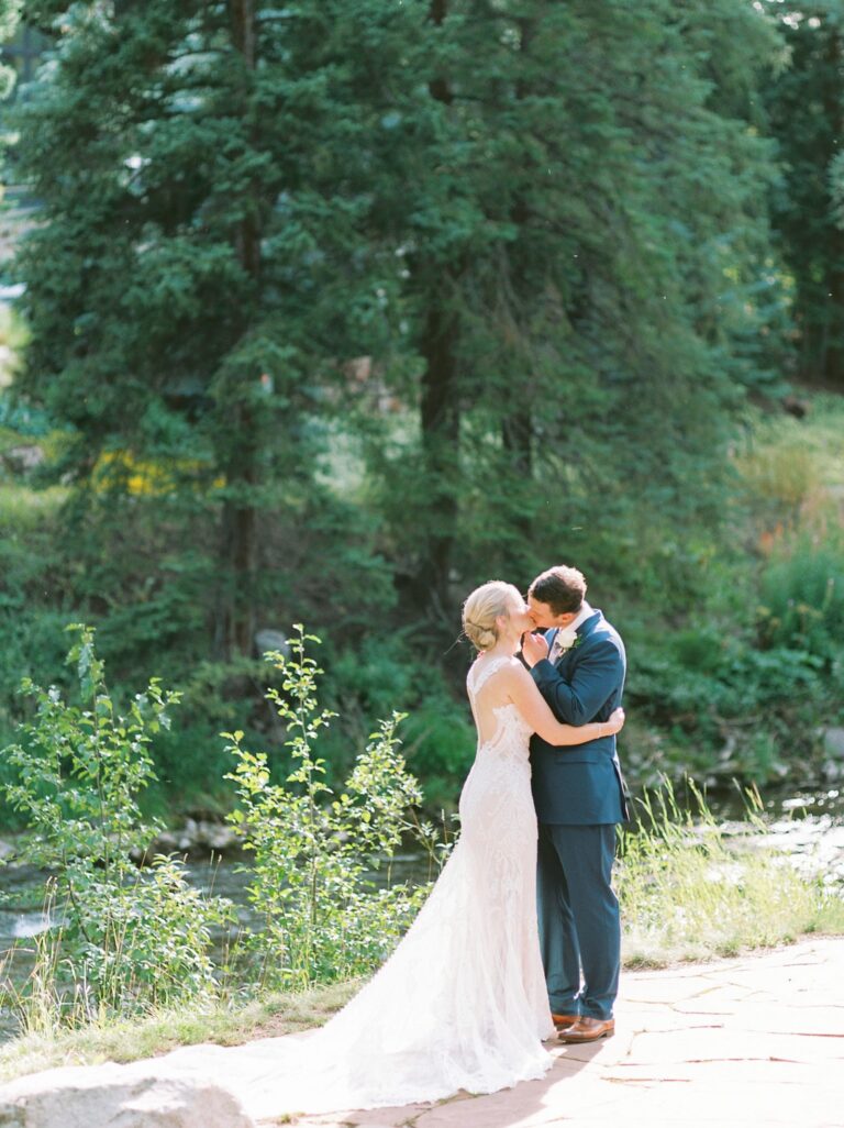Bride and groom standing outdoors on a sunny day, next to a stream, kissing