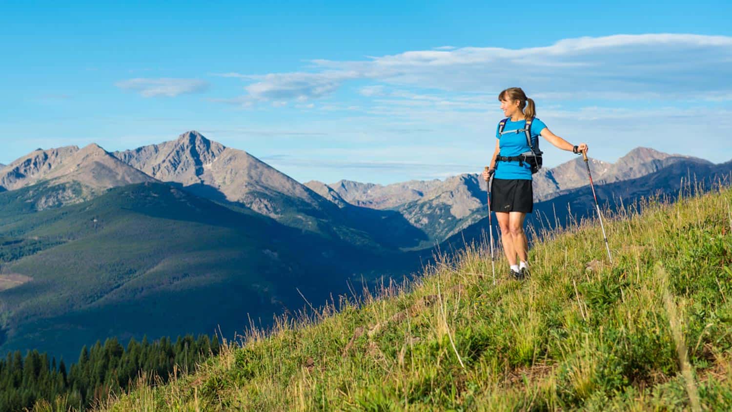 Woman hiking outdoors, holding poles, looking over at valley and mountains in the distance