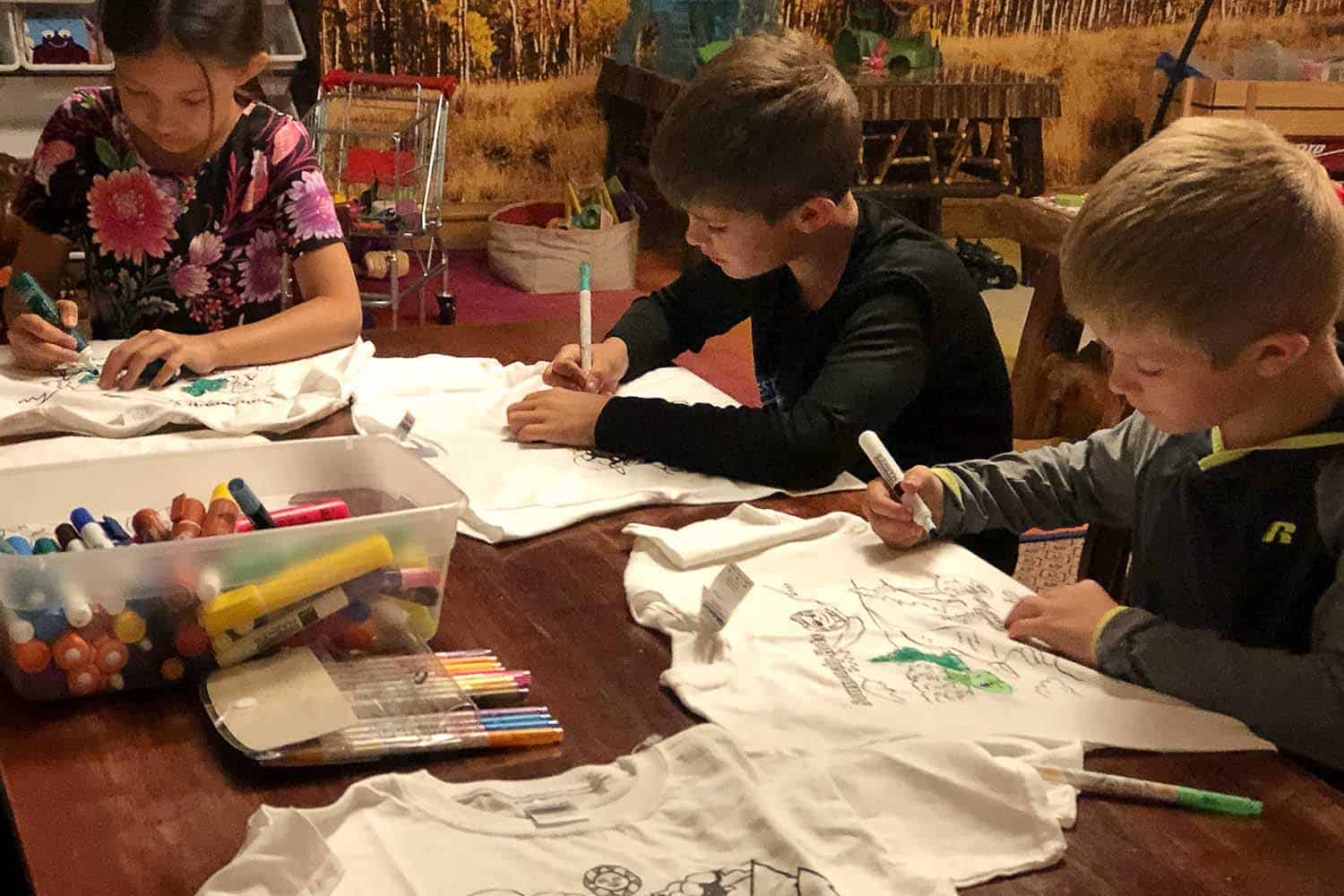 Young childresn seated at table, painting shirts with markers