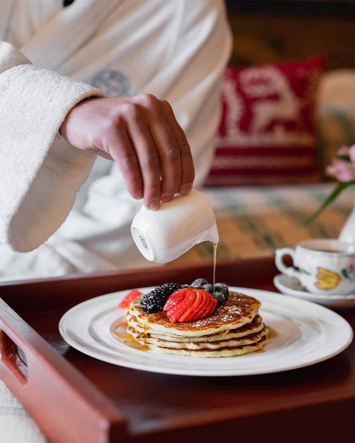 Person wearing bathroube pouring syrup over pancakes with fruit on tray on bed