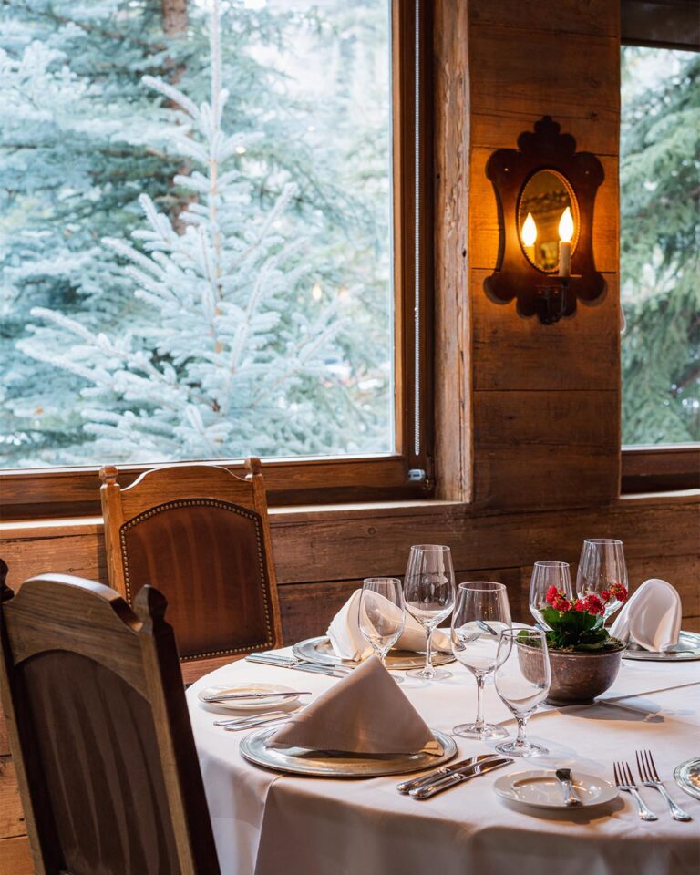Round table set for dinner, chairs, next to window with view of snow-covered pine trees in the background