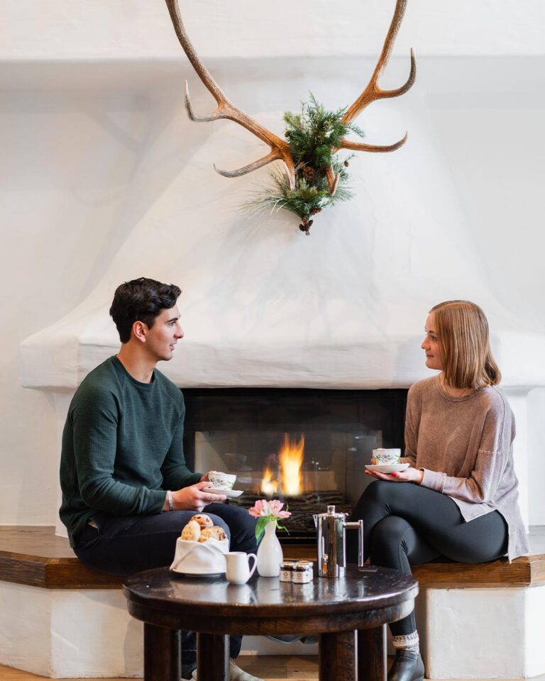 Couple holding cups, sitting next to fireplace with antlers on the wall above, and table with pastries and kettle in the foreground