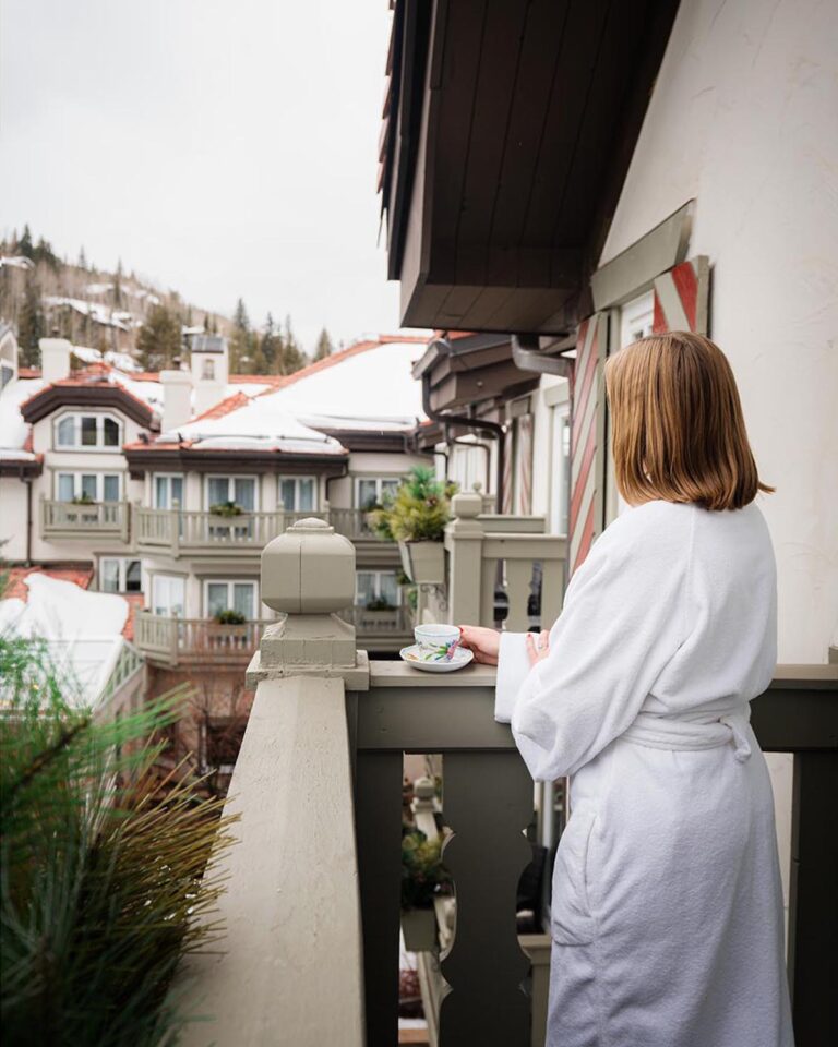Woman wearing bathrobe, on a balcony, holding cup of coffee and looking over at snow-covered rooftops