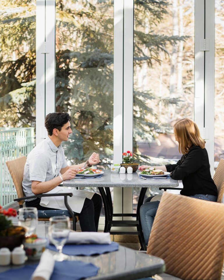 Couple enjoying a meal, sitting at covered outdoor restaurant area with large glass doors with view of trees