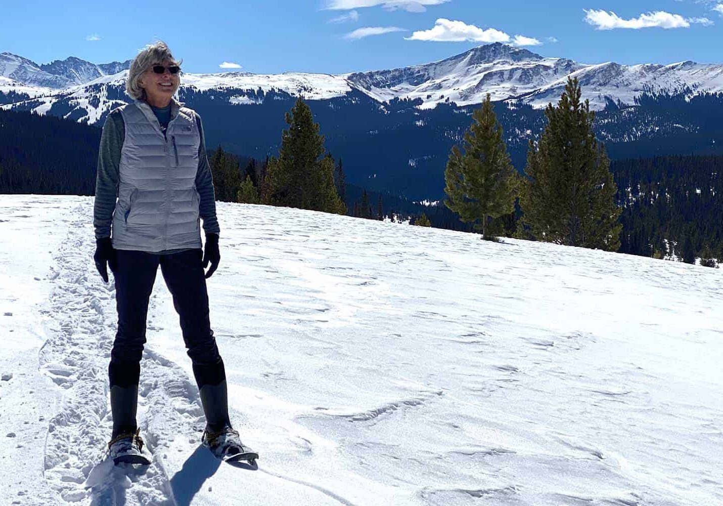 Woman wearing vest, snow shows, and snow covered mountains in the background on a sunny day