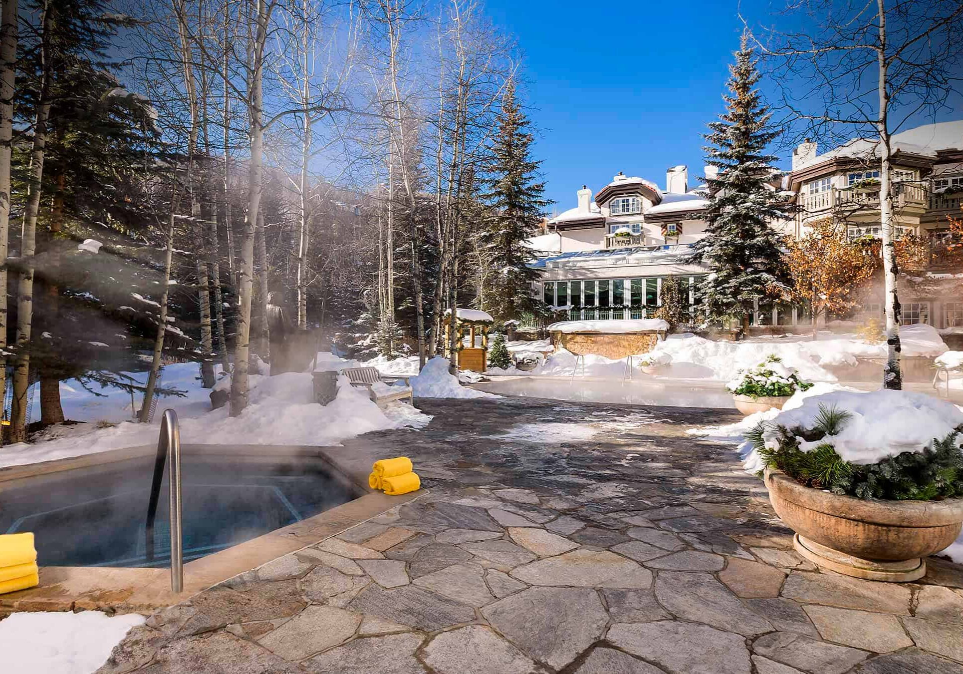 Steam rises from hot tub on a sunny day, as snow covers the ground and spa building is visible in the background