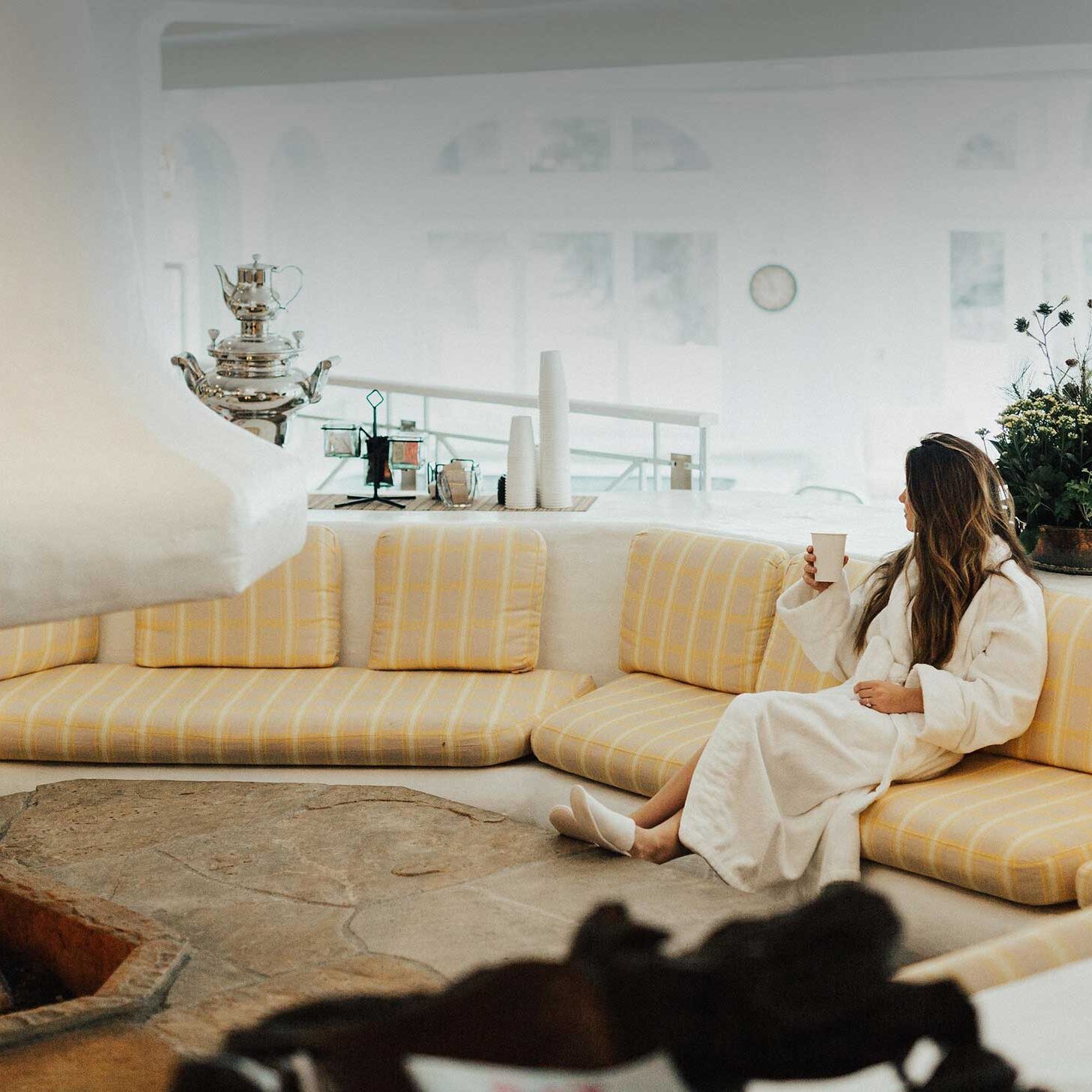 Woman wearing robe and slippers, drink in hand, sitting on cushions