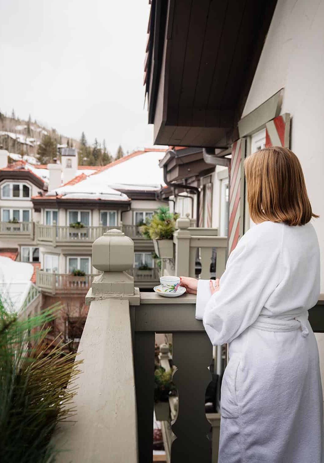 Woman wearing bathrobe, on a balcony, holding cup of coffee and looking over at snow-covered rooftops